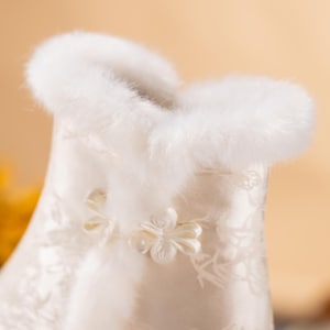 Peut inclure: Un v&ecirc;tement en satin blanc orn&eacute; d'un motif floral d&eacute;licat et d'une bordure &eacute;paisse en fausse fourrure blanche douce le long du col. La pi&egrave;ce est ferm&eacute;e par un petit bouton en tissu en forme de fleur complexe au niveau du cou.