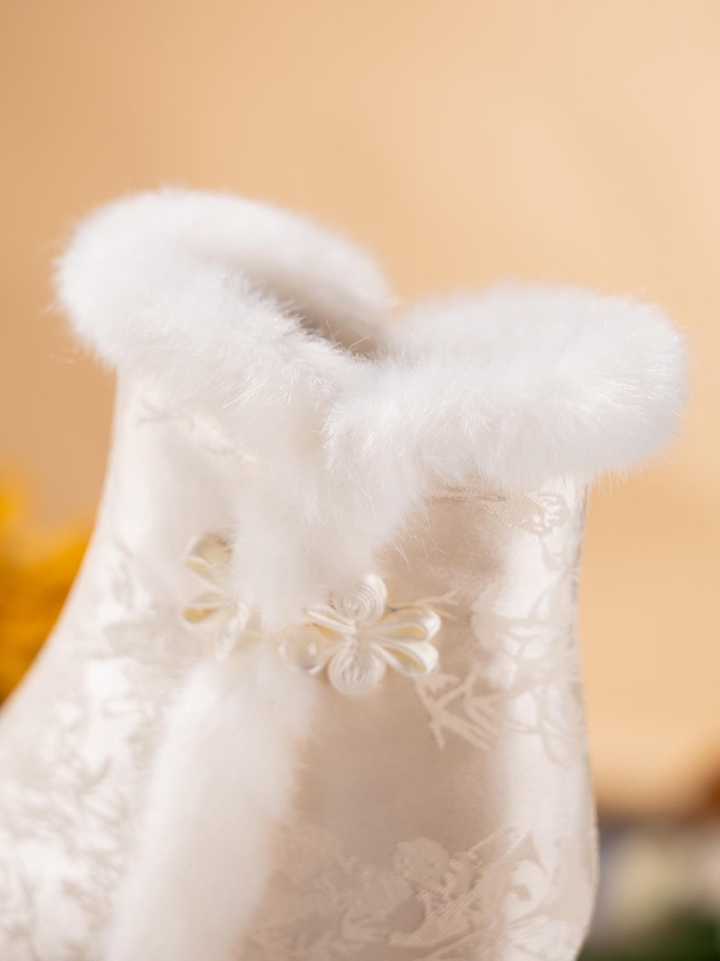 Peut inclure: Un v&ecirc;tement en satin blanc orn&eacute; d'un motif floral d&eacute;licat et d'une bordure &eacute;paisse en fausse fourrure blanche douce le long du col. La pi&egrave;ce est ferm&eacute;e par un petit bouton en tissu en forme de fleur complexe au niveau du cou.