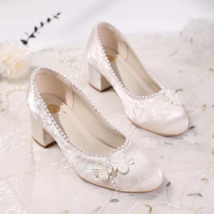 May include: A pair of ivory-colored block heel shoes. The shoes feature a floral pattern, pearl trim, and a decorative flower on the toe. The heels are approximately 2 inches high.