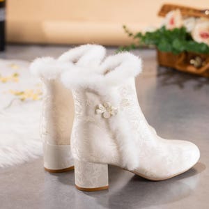 Peut inclure: Une paire de bottines blanc cass&eacute; avec un motif botanique ton sur ton subtil, une bordure &eacute;paisse en fausse fourrure blanche autour du col et de la couture lat&eacute;rale, et une petite d&eacute;coration florale blanche sur le c&ocirc;t&eacute;. Talon bloc et bout rond.