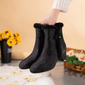 Peut inclure: Bottines noires avec un motif floral ton sur ton discret, une bordure en fausse fourrure noire le long de l'ouverture sup&eacute;rieure, un talon bloc et une fermeture d&eacute;corative en tissu noir avec une petite perle ronde vert vif sur le c&ocirc;t&eacute;.
