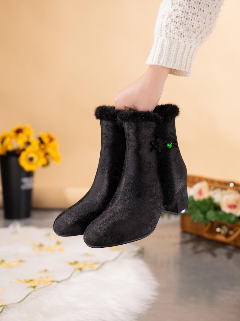 Peut inclure: Bottines noires avec un motif floral ton sur ton discret, une bordure en fausse fourrure noire le long de l'ouverture sup&eacute;rieure, un talon bloc et une fermeture d&eacute;corative en tissu noir avec une petite perle ronde vert vif sur le c&ocirc;t&eacute;.
