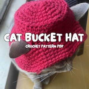 Può includere: Un cappello a secchiello rosso all'uncinetto con orecchie da gatto, indossato da un gatto. Il cappello presenta la scritta bianca "CAT BUCKET HAT" e "CROCHET PATTERN PDF". Le orecchie del gatto sono visibili, aggiungendo un tocco giocoso.