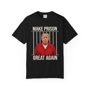 May include: A black short-sleeve t-shirt featuring a graphic of a person in a red prison uniform behind grey vertical bars. The text "MAKE PRISON" appears above the bars and "GREAT AGAIN" appears below in a light-colored, sans-serif font.