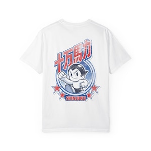 Könnte beinhalten: Ein weißes Kurzarm-T-Shirt mit einem Grafikdruck auf der Rückseite, der die Figur Astro Boy in einem kreisförmigen Rahmen mit roten Sternen, dem japanischen Text 十万馬力 und dem Wort SHINJUKU in roten Buchstaben zeigt, alles mit Used-Optik.