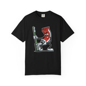 Op de afbeelding: Een zwart t-shirt met korte mouwen en een grafische print van een cartoon-tandpastatube met een gezicht, armen en benen. Het personage knijpt witte tandpasta op een groene tandenborstel. Het ontwerp bevat rode, witte en groene kleuren.