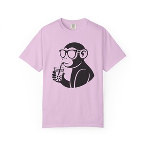Puede incluir: Una camiseta de manga corta de color morado claro con un estampado gráfico negro de un mono con gafas que bebe de un vaso con pajita. El mono aparece de perfil, sosteniendo un recipiente de bebida lleno de perlas de boba redondas.