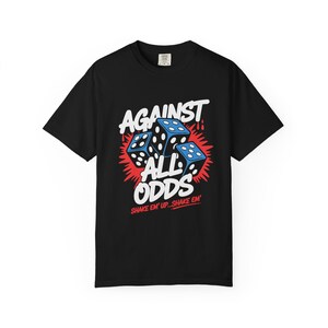 May include: A black short-sleeve t-shirt featuring a graphic print of three blue dice with white dots. The text reads AGAINST ALL ODDS in large white letters, with the red text SHAKE EM' UP...SHAKE EM' positioned below the dice illustration.