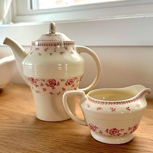 May include: Cream-colored ceramic teapot and creamer set. The teapot has a lid and a curved spout, and the creamer has a handle. Both pieces are decorated with a repeating floral pattern in a deep red color. The set is placed on a wooden surface.