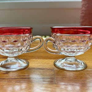 May include: Two clear glass teacups with a red rim and a diamond pattern. The teacups have a handle and sit on a clear, pedestal base. The teacups are on a wooden surface.