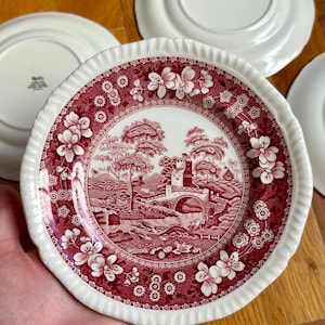 May include: A round ceramic plate featuring a deep red transferware pattern of a stone bridge, a castle, and trees. The rim has a scalloped edge and a floral border. The back of the plate is white with a black stamp that reads Spode England.