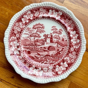 May include: A round, decorative plate with a red and white floral and landscape design. The plate features a scalloped edge and a detailed illustration of a bridge, trees, and a castle. The plate is made of ceramic.