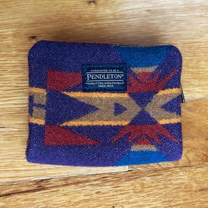 May include: A rectangular zippered pouch featuring a multi-colored geometric pattern in shades of purple, blue, red, and tan. A dark blue rectangular fabric label is centered on the front with the white text: WARRANTED TO BE A PENDLETON PENDLETON WOOLEN MILLS SINCE 1863.