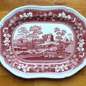 May include: An oval serving platter with a red and white floral and landscape design. The platter features a detailed pastoral scene in the center, surrounded by a floral border and a scalloped edge. The color palette is primarily red and white.