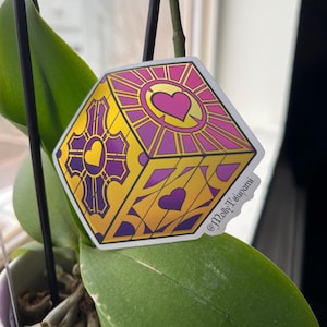 May include: A sticker featuring a geometric design of a yellow, purple, and gold box with heart shapes. The sticker has the text "@MollyTsunami" in the bottom right corner. The sticker is placed in front of a green plant.