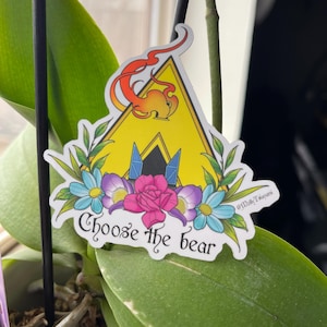 May include: A sticker with a yellow triangle design, a flame, and the text "Choose the bear." The sticker is decorated with blue, purple, and pink flowers and green leaves. The sticker is on a green leaf.