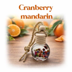 May include: A clear glass car air freshener with a wooden cap and a decorative rope. The freshener contains colorful dried flowers and the text "Cranberry mandarin" in orange. Oranges are in the background.