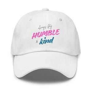 May include: A white baseball cap featuring the text "always stay" in black script, "HUMBLE" in a bold pink brushstroke font, and "& kind" in a bold blue brushstroke font centered on the front panel of the hat with a curved brim.