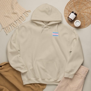May include: A sand-colored hooded sweatshirt featuring a small rectangular transgender pride flag patch on the left chest. The hoodie includes a front kangaroo pocket, long sleeves, and matching drawstring cords at the neckline for an adjustable fit.