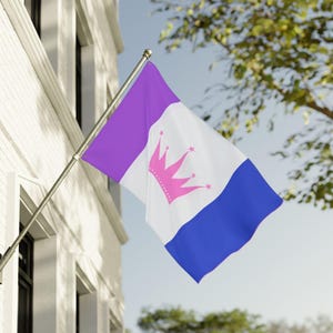 May include: A rectangular flag attached to a metal pole, featuring three vertical stripes in purple, white, and blue. The centre white stripe displays a pink crown graphic with small star accents at the tips, waving against a bright sky.