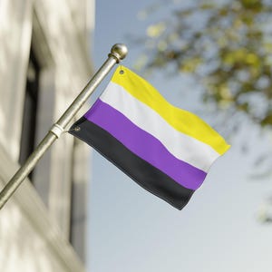 May include: A non-binary pride flag waving in the breeze. The flag features horizontal stripes in black, purple, white, and yellow. The flag is attached to a silver pole with a gold finial, set against a building and a blue sky.