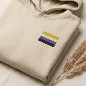 May include: A beige hooded sweatshirt featuring an embroidered non-binary pride flag on the chest. The flag consists of four horizontal stripes in yellow, white, purple, and black. The hoodie has a drawstring hood and a front kangaroo pocket.