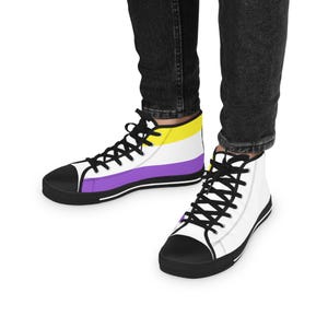 May include: High-top trainers with a black sole, black laces, and a white upper. The trainers feature a non-binary pride flag design with yellow, white, and purple stripes. The shoes are worn with black jeans.