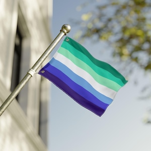 May include: A flag with horizontal stripes of dark purple, blue, light blue, white, green, and teal, waving in the wind. The flag is attached to a silver pole with a gold ball finial. The background includes a building and a blurred view of trees.
