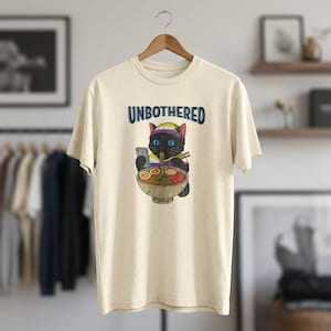 Peut inclure: T-shirt crème avec le mot "UNBOTHERED" au-dessus d'un chat de dessin animé portant un bandeau et tenant un téléphone tout en mangeant des ramen. Le chat est noir avec des yeux bleus. Le bol de ramen contient des nouilles, des œufs et de la viande.