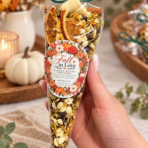 May include: A clear cone-shaped bag filled with a potpourri simmer blend of dried orange slices, cinnamon sticks, cloves, apple pieces, calendula petals, and jasmine flowers. A label reads Fall in Love Simmer Blend, Net Wt. 2.0 oz.