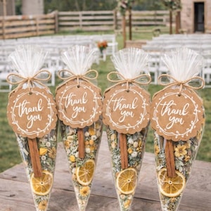 May include: Four clear cone-shaped bags filled with dried flowers, spices, and a slice of dried orange. Each bag has a brown tag with the words "Thank you" in white script, tied with twine. The bags are arranged on a wooden surface, with a blurred outdoor background.