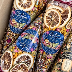 May include: Three cone-shaped clear plastic bags filled with dried botanicals, including orange slices, cinnamon sticks, and flower petals. Each bag features a blue label reading New Year, NEW GOALS SIMMER BLEND and a red string tie at the top.