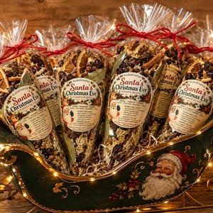 May include: Five clear plastic cone-shaped bags filled with a dried Santa's Christmas Eve simmer blend of orange slices, cranberries, and spices. Each bag has a label with a Santa illustration and red twine tie, displayed in a green metal sleigh.