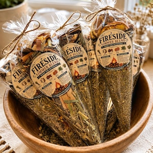May include: Several cone-shaped clear plastic bags filled with dried botanicals, including orange slices and cinnamon sticks, labeled Fireside Simmer Blend Made for Outdoors. Each bag is tied with brown twine at the top and contains a mix of herbs.