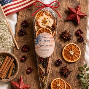 May include: A clear cone-shaped bag filled with dried orange slices, cinnamon sticks, star anise, and berries. A round label reads Stars, Stripes & Simmers, The All-American Simmer Blend. The bag is tied with a red string at the top.
