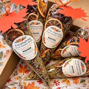 May include: Four clear cone-shaped bags filled with a Friendsgiving Feast Simmer Blend of dried orange slices, cranberries, rosemary, and cinnamon sticks. Each bag is tied with orange twine and features a red paper maple leaf tag.