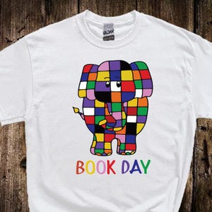 Könnte beinhalten: Weißes T-Shirt mit einem farbenfrohen Patchwork-Elefanten-Design. Der Elefant besteht aus Quadraten in verschiedenen Farben, darunter Rot, Blau, Grün und Gelb. Unter dem Elefanten sind die Worte "BOOK DAY" in Regenbogenfarben gedruckt.