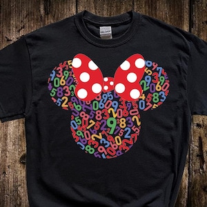 May include: Black t-shirt featuring a Minnie Mouse design. The design includes a red bow with white polka dots and a mouse head shape filled with colorful numbers. The shirt is made of a soft material.