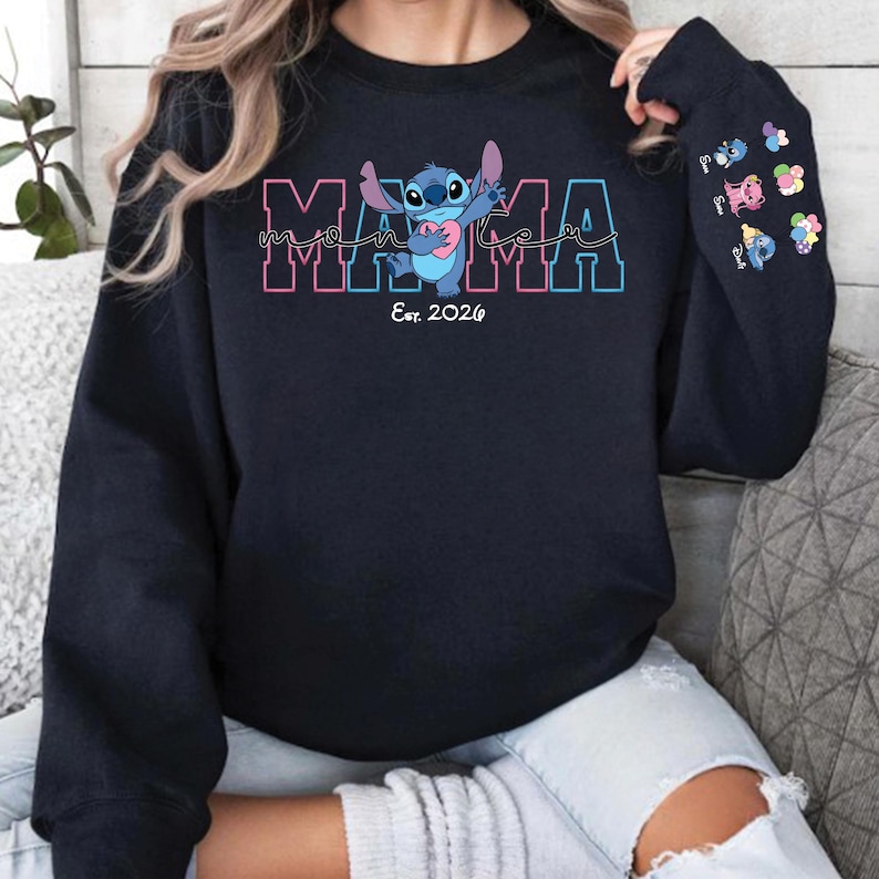 Puede incluir: Una sudadera azul marino con la palabra "MAMA" en rosa y azul, y el personaje Stitch sosteniendo un coraz&oacute;n. El texto "Est. 2026" est&aacute; debajo de la palabra "MAMA". La manga tiene varios gr&aacute;ficos de personajes peque&ntilde;os.