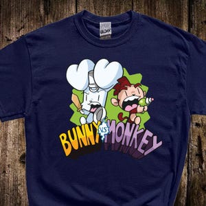May include: Navy blue t-shirt featuring a cartoon graphic of a bunny holding a bat and a monkey holding a green object. The text "BUNNY vs MONKEY" is in yellow and purple.