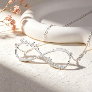 May include: A silver-toned infinity symbol necklace featuring the names Sophia and Mason integrated into the design. The pendant is attached to a delicate silver-toned chain, showcasing a polished metallic finish and cursive lettering.