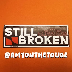 May include: A rectangular sticker with the words "STILL BROKEN" in large white letters. The word "BROKEN" has a red and black design. Above the word "BROKEN" is Japanese text and a red graphic. The sticker also includes the text "@AMYONTHETOUGE".
