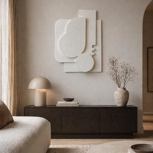 Pode incluir: Uma sala de estar moderna com um armário de madeira castanho escuro, um sofá bege e uma mesa de centro redonda. Uma lâmpada bege e um vaso texturizado com flores secas estão no armário. Uma escultura de parede abstrata branca adiciona um toque minimalista.