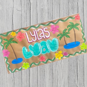 May include: A hand-drawn banner with the text "Lyla's Way" in bubble letters. The banner features palm trees, hibiscus flowers, and a blue ocean scene, all outlined in green and set against a tan background.