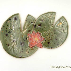 May include: Handmade ceramic lily pad and flower set. The lily pads are a mottled green with dark speckles, and the flower is a vibrant red with yellow accents. The set is designed to be a decorative piece.