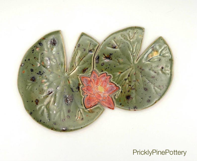 May include: Handmade ceramic lily pad and flower set. The lily pads are a mottled green with dark speckles, and the flower is a vibrant red with yellow accents. The set is designed to be a decorative piece.