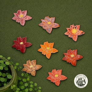 May include: Eight ceramic lotus flower ornaments in various shades of pink, red, orange, and brown, each featuring a yellow center. A circular white logo with a black hedgehog illustration and the text Prickly Pine Pottery is in the corner.