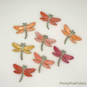 May include: A collection of handmade ceramic dragonfly ornaments in various colors, including orange, yellow, and red. Each dragonfly has detailed wings and a textured body, perfect for home decor or craft projects.