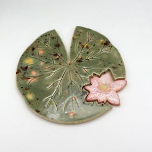 以下が含まれることがあります： 薄緑色のベースとピンクの花のアクセントが付いた、セラミック製のスイレンの葉の形をした皿。 皿には、葉の脈のパターンと、茶色と黄色の斑点模様が施されています。 花は、赤い輪郭のソフトピンクです。