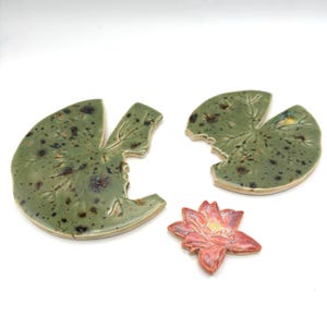 May include: Ceramic lily pad and lotus flower set. The lily pads are green with brown speckles and leaf vein details. The lotus flower is pink with yellow and orange accents.